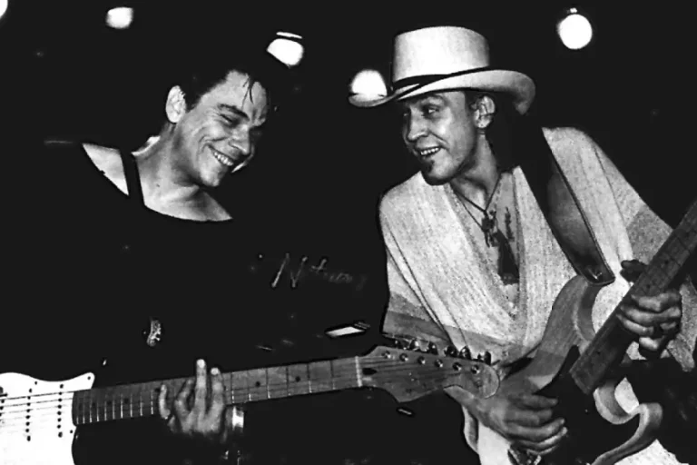 Stevie Ray Vaughan and Jimmie Vaughan perform side by side onstage, exchanging smiles while playing guitar, capturing their musical chemistry in a live setting.