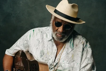 Portrait of Taj Mahal holding an acoustic guitar, wearing a light-colored hat and sunglasses, conveying a reflective, soulful mood.