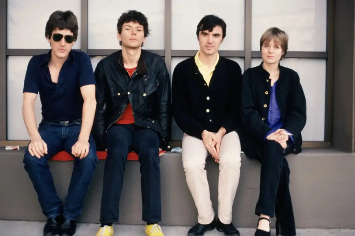 Talking Heads sit together casually on a ledge outdoors, capturing the band’s relaxed, modern look during their later years.