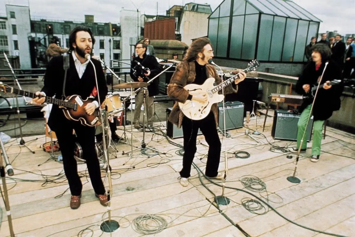 The Beatles performs on a rooftop, with two guitarists singing into microphones while a drummer plays behind them.