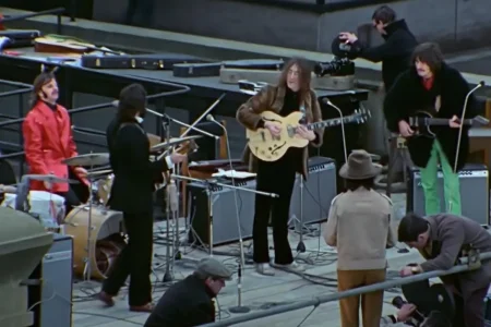 The Beatles music performance in progress, showing multiple musicians with electric guitars and drums as a camera operator films them at close range.