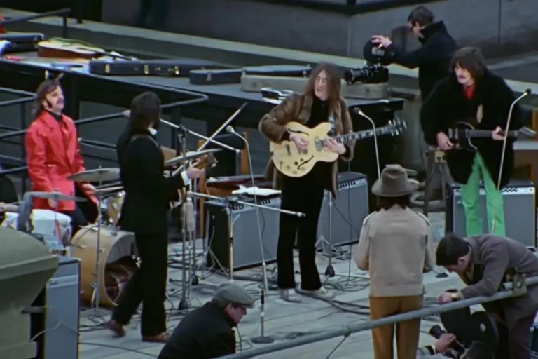 The Beatles music performance in progress, showing multiple musicians with electric guitars and drums as a camera operator films them at close range.