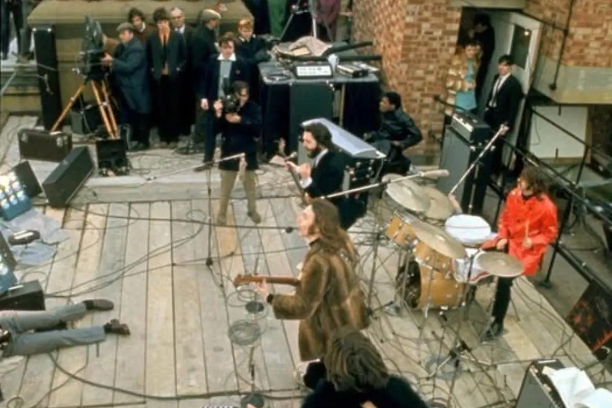 The Beatles with a full band playing among microphones, amplifiers, and tangled cables while a small crowd and film crew watch nearby.