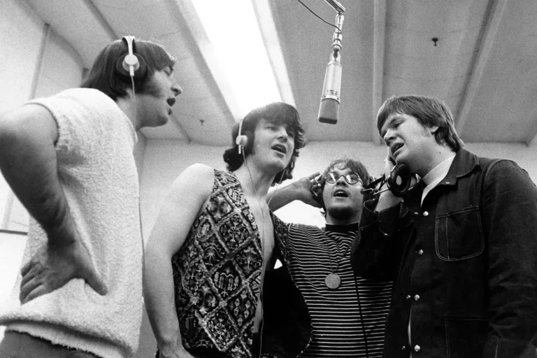 The Guess Who singing together in a recording studio, wearing headphones around a shared microphone during a vocal session.