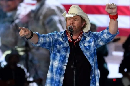 Toby Keith performs live onstage in a blue plaid shirt and white cowboy hat, pointing toward the crowd while singing into a microphone with an American flag in the background.