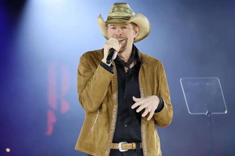 Toby Keith smiles while holding a microphone on stage, dressed in a tan jacket and cowboy hat against a softly lit concert backdrop.