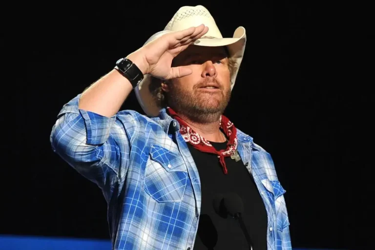 Toby Keith’s “American Soldier”: The Backstory, the Backlash, the Legacy Toby Keith wearing a white cowboy hat, blue plaid shirt, and red bandana, raising a hand to his brow under stage lighting.