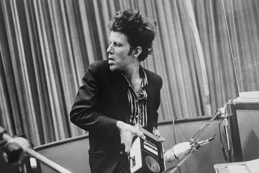 Tom Waits in a recording studio holding a reel-to-reel tape box, dressed in a dark jacket, photographed in black and white.
