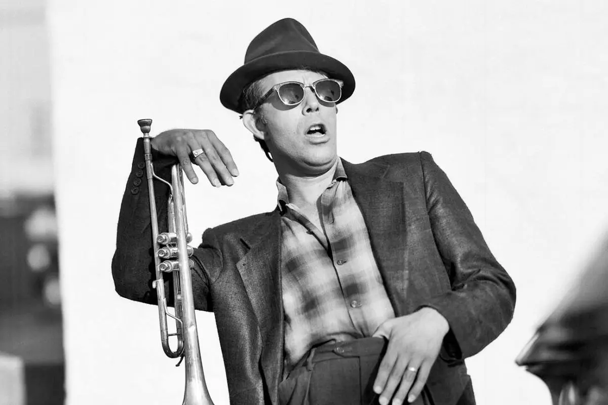 Tom Waits wearing a hat and sunglasses, holding a trumpet in a black-and-white photograph.
