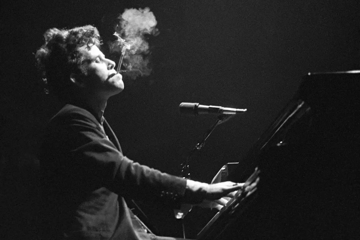 Tom Waits performs at a piano on stage, cigarette smoke drifting upward, capturing the raw, late-night intensity of his live performances.