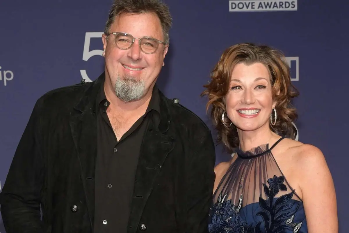 Vince Gill and Amy Grant smiling while holding Grammy Awards at an awards event, standing in front of a step-and-repeat backdrop.