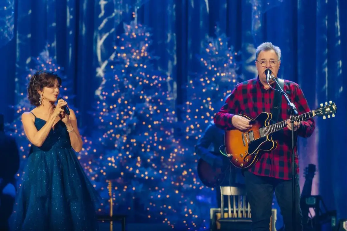 Amy Grant singing into a microphone while Vince Gill plays guitar and sings onstage, with softly lit Christmas trees in the background.
