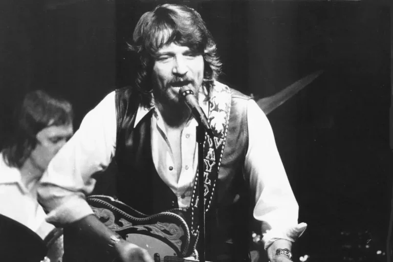 Waylon Jennings performing on stage, singing into a microphone while playing an acoustic guitar, wearing a vest over a button-down shirt.