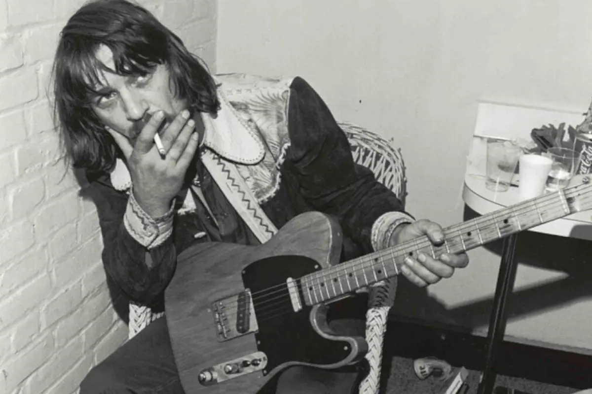 Waylon Jennings seated indoors, holding an electric guitar and a cigarette, looking directly at the camera with a relaxed expression.