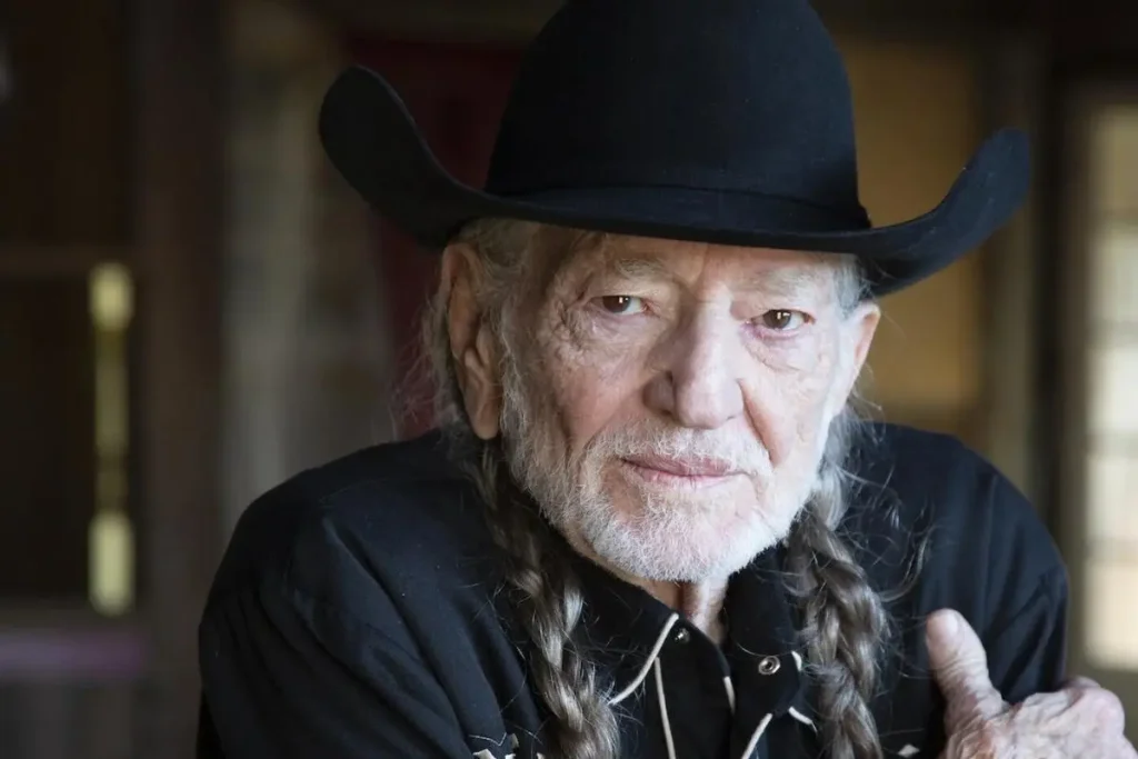 Willie Nelson wearing a black cowboy hat with long braided hair, looking directly at the camera in a softly lit indoor setting.