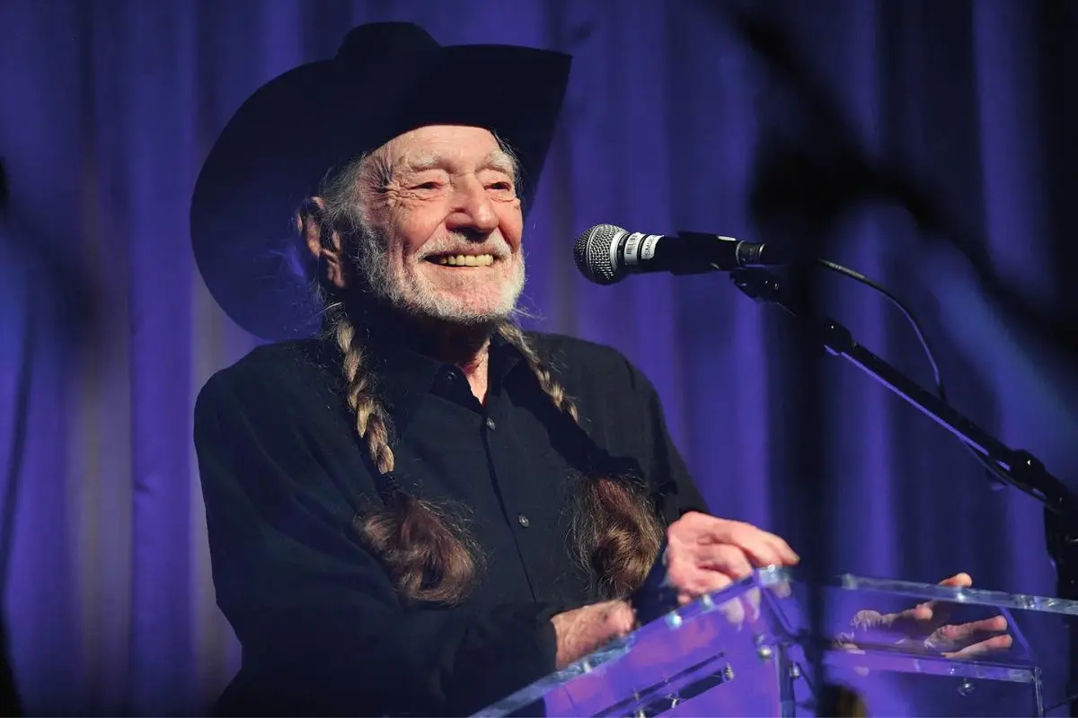 Willie Nelson smiling while standing at a microphone on stage, wearing a black shirt and cowboy hat under blue stage lighting.