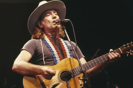Willie Nelson singing into a microphone while playing acoustic guitar.
