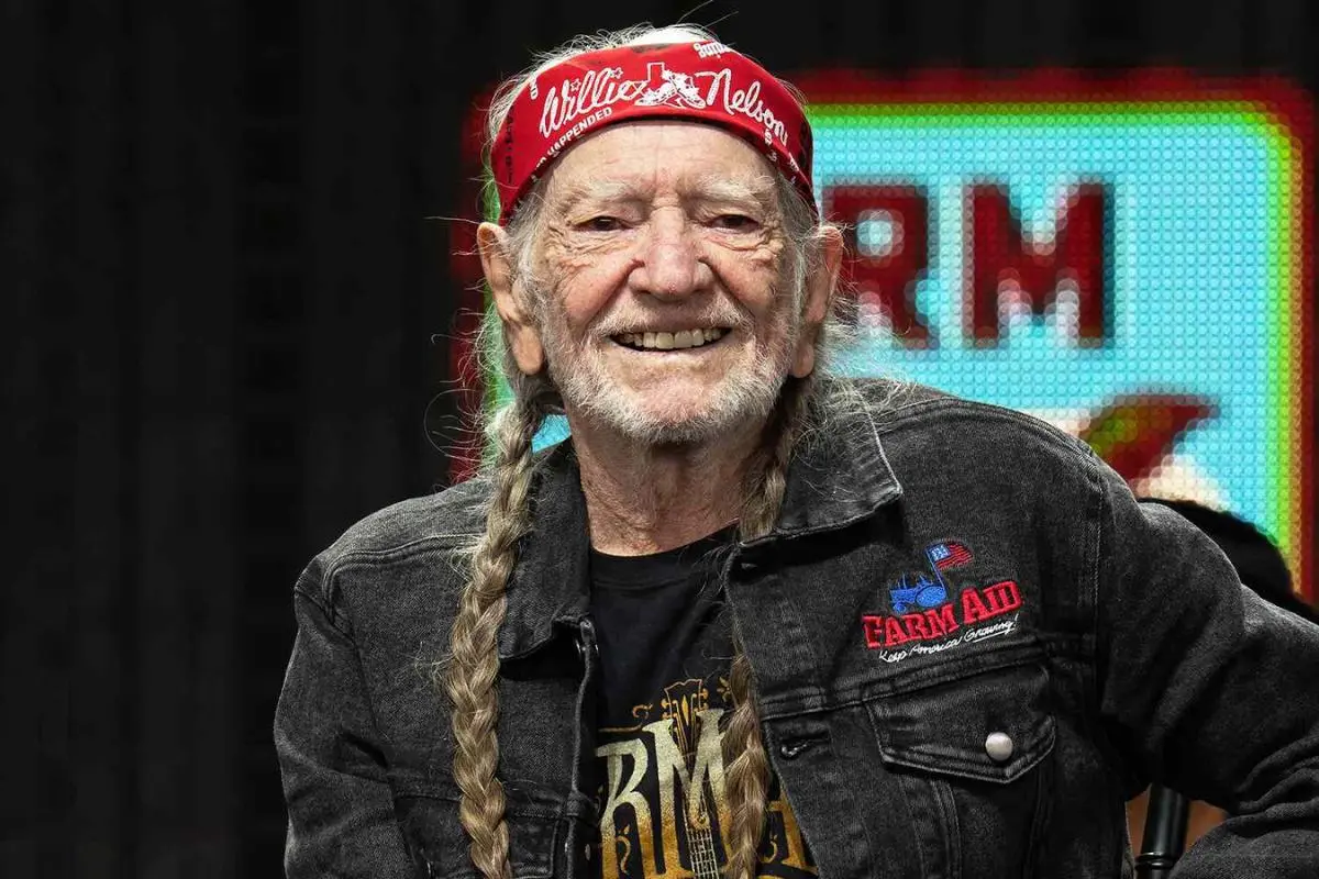 Willie Nelson smiling on stage, wearing a red bandana, long braids, and a dark jacket with embroidered patches.