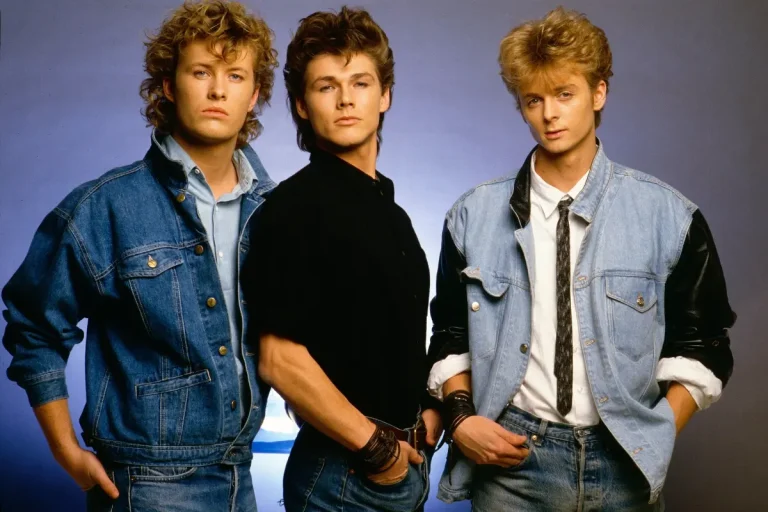 A-ha band posed in classic mid-’80s fashion: denim, sharp silhouettes, and carefully styled hair, photographed against a studio backdrop.