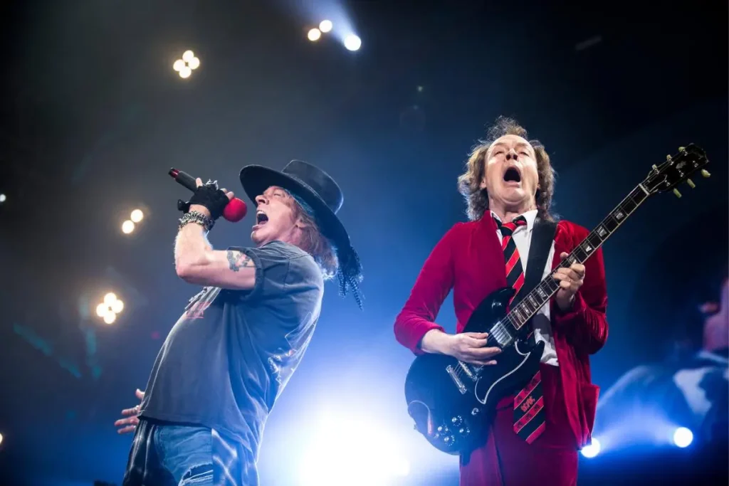 ACDC sing and play electric guitar side by side on a brightly lit concert stage.
