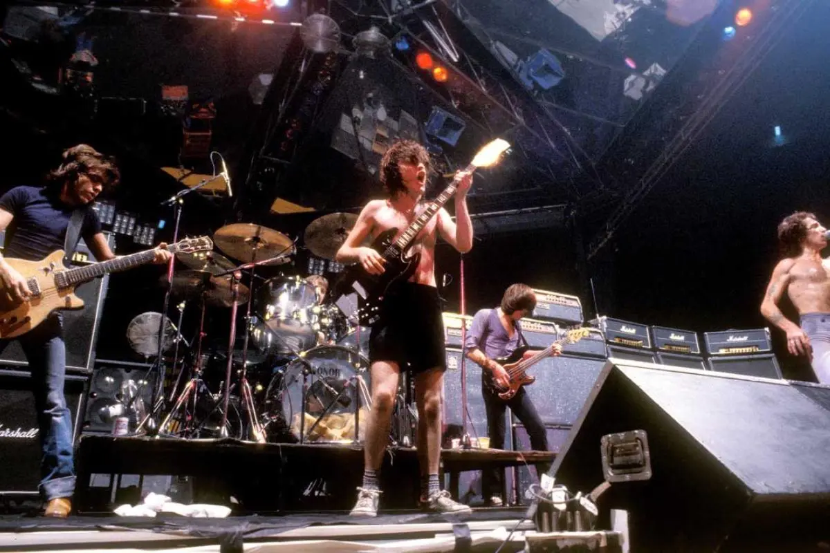 ACDC performs live on stage, with a guitarist in the center playing energetically while other band members play guitar and bass near large amplifiers and a drum kit.