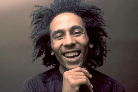 Bob Marley laughing, head tilted slightly back, hair loose and frizzy, showing a wide toothy smile.