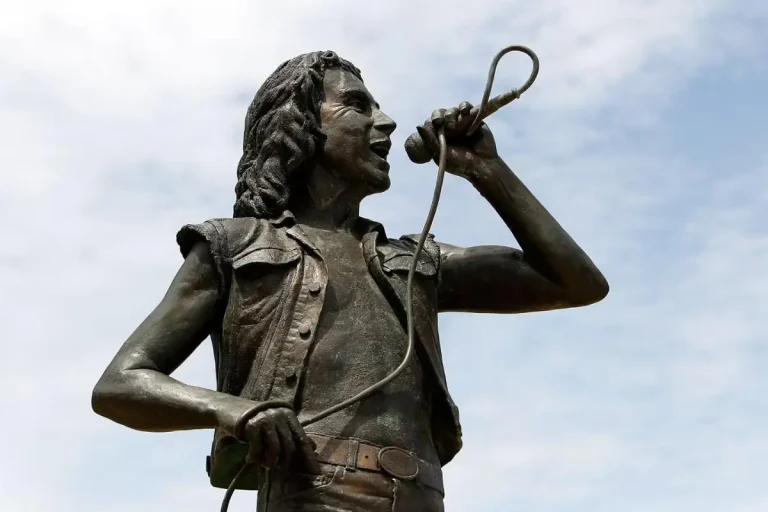 Bon Scott statue singing into a microphone, capturing the detailed facial expression, sleeveless vest, and muscular pose against the sky.