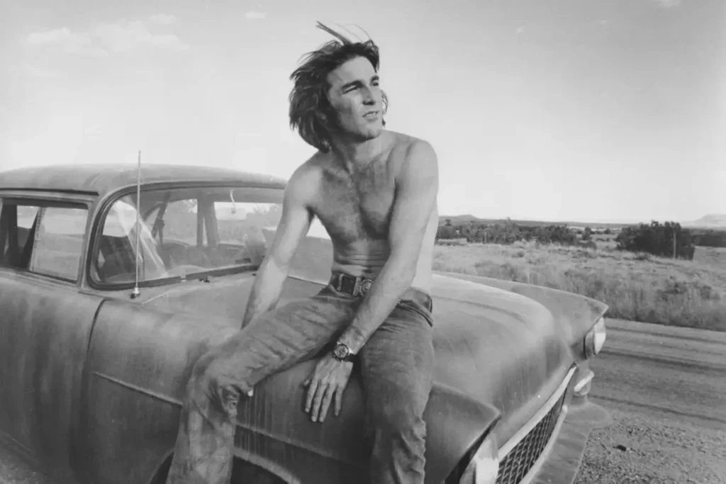 Dennis Wilson: The Only Beach Boy Who Actually Surfed (And Lived the Myth) Dennis Wilson sitting on the hood of a dusty vintage car in a rural landscape, looking off into the distance.