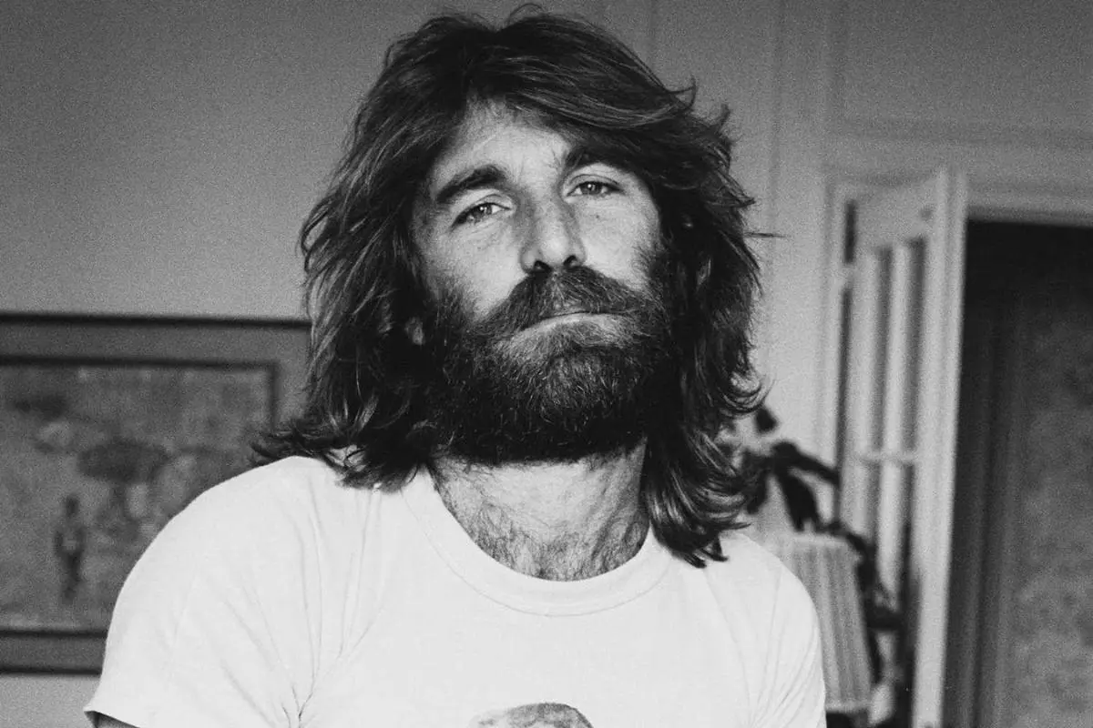 Dennis Wilson wearing a light-colored T-shirt indoors, gazing calmly at the camera with a relaxed expression.