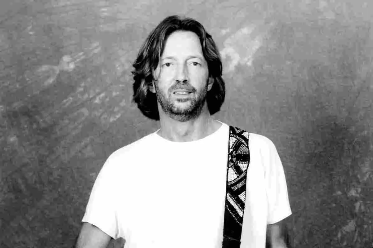 Eric Clapton standing alone, facing the camera with a guitar strap over his shoulder.