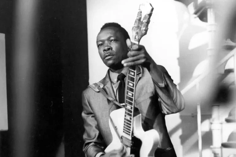 John Lee Hooker holding an electric guitar onstage, captured mid-performance with a focused, commanding expression.