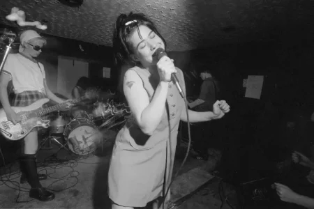Kathleen Hanna performing in an intimate club setting, gripping a microphone while bandmates play behind her, capturing raw DIY energy.