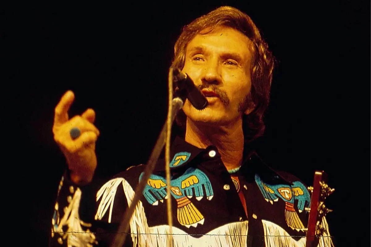 Marty Robbins performing on stage under warm lighting, wearing a black western shirt with colorful embroidered birds.