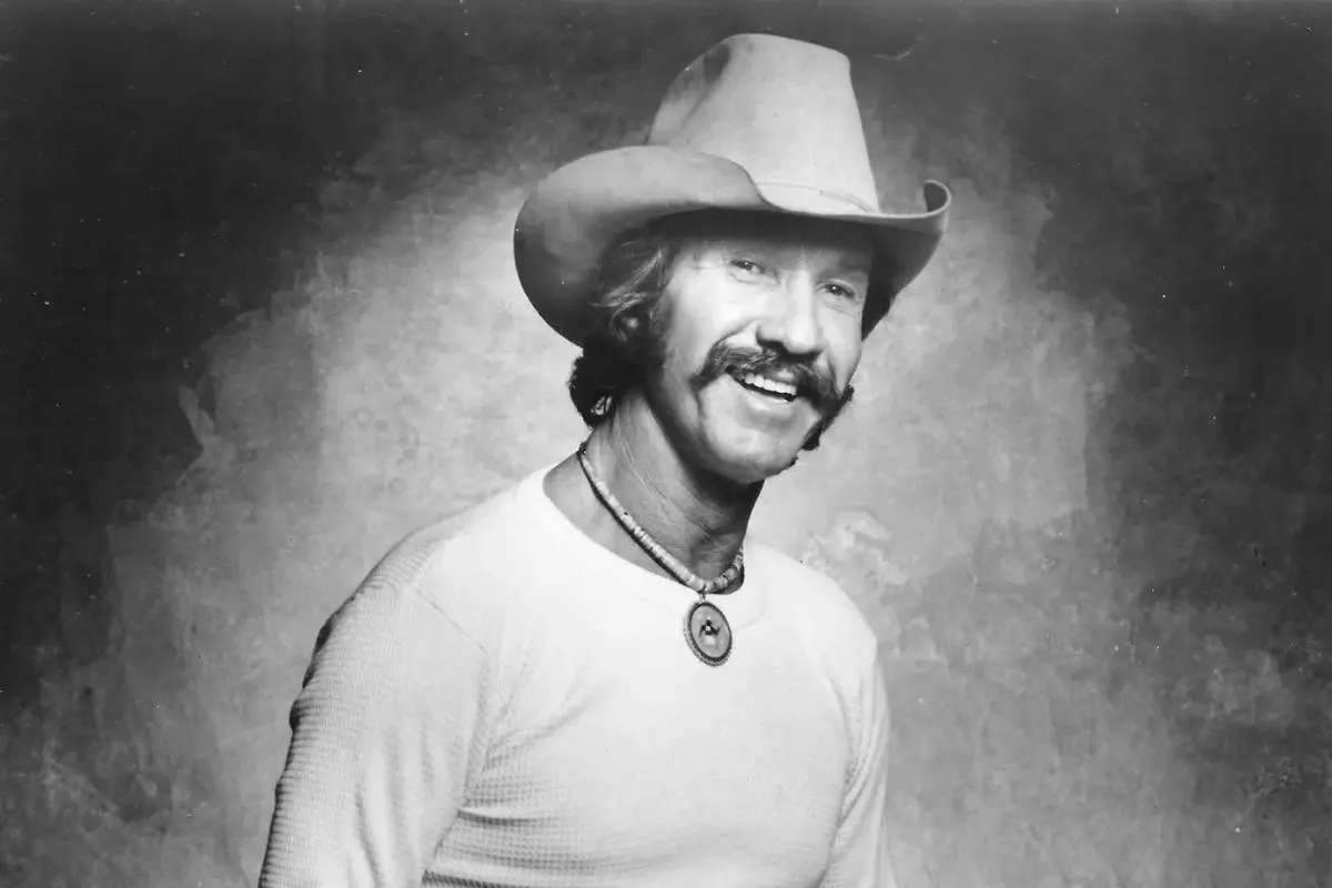 Marty Robbins smiling country singer wearing a cowboy hat and a light-colored long-sleeve shirt, standing against a textured studio backdrop.