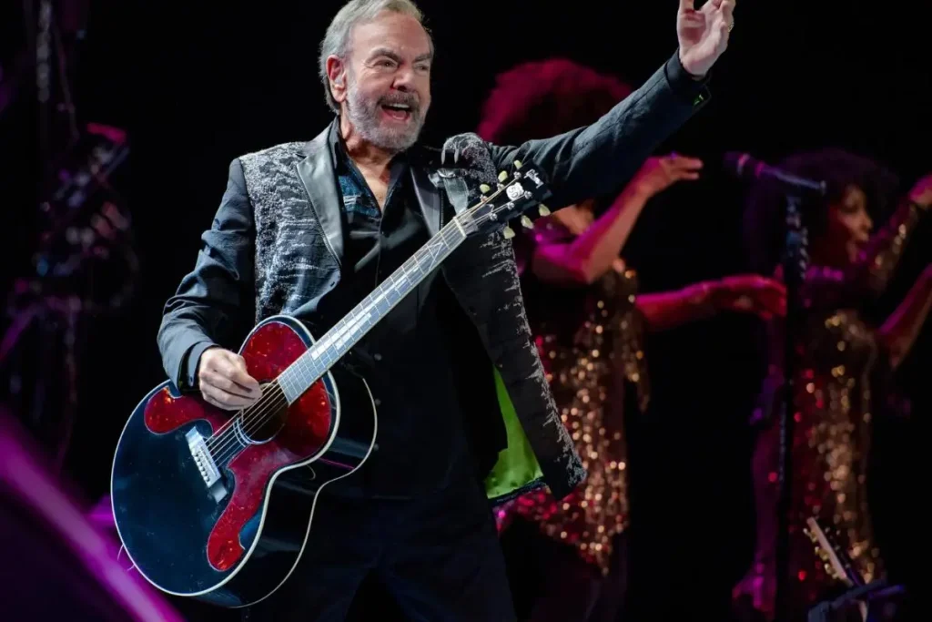 Neil Diamond smiles and raises one arm while holding an acoustic guitar onstage.