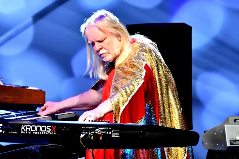 Rick Wakeman playing a keyboard onstage, wearing a flowing cape under colorful stage lighting during a concert performance.
