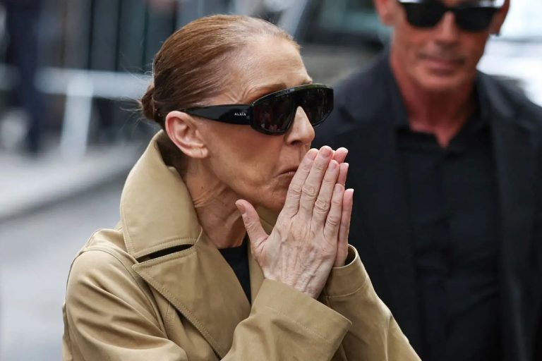 Celine Dion in a camel trench coat and dark sunglasses presses her hands together and blows a kiss to fans on a Paris street