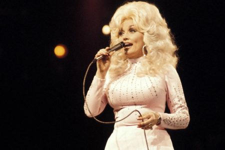 Dolly Parton performing on stage in the 1970s, singing into a handheld microphone and wearing a pale pink rhinestone-studded dress