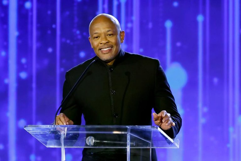 Dr. Dre Says the Only People Who Fear AI Are Those Who ‘Have Trouble Creating’ Dr. Dre speaking at a clear acrylic podium against a blue lit backdrop, smiling with both hands raised