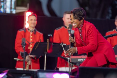 Gladys Knight performing in a red suit, holding a microphone and smiling, with United States Marine Band musicians in red dress uniforms behind her