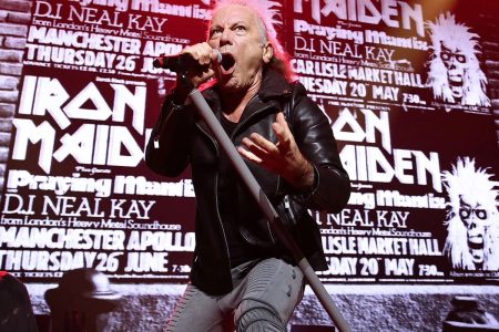 Iron Maiden singer Bruce Dickinson sings into a handheld microphone on stage in a black leather jacket, in front of a large screen showing vintage black-and-white Iron Maiden concert posters