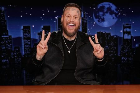 Jelly Roll smiling and holding up two peace signs behind the host desk on Jimmy Kimmel Live, with the show's night-time city skyline backdrop behind him