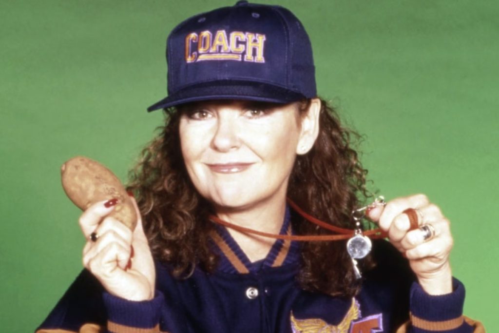 Shelley Fabares in a navy Coach team jacket and cap, smiling and holding up a potato in one hand and a whistle in the other, against a green backdrop