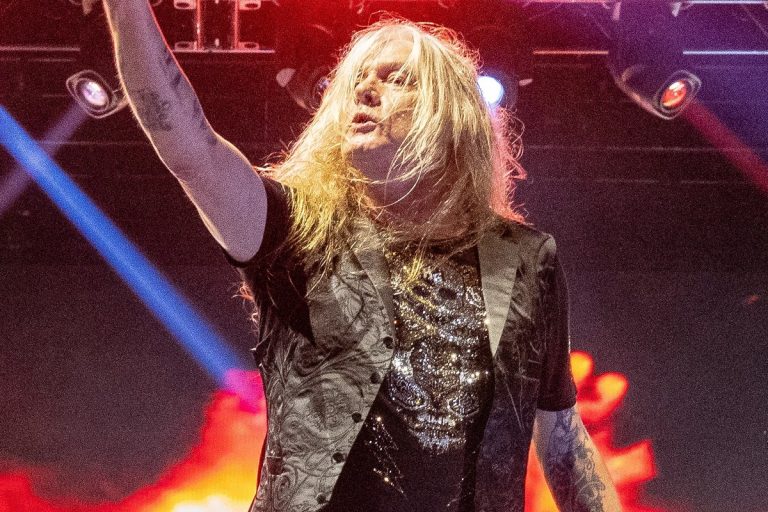 Sebastian Bach Fronts Twisted Sister for the First Time as Dee Snider’s Health Keeps Him Off the Road Sebastian Bach singing on stage with one arm raised, lit by red and blue stage lights