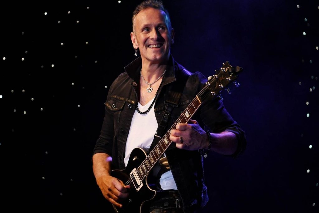 Def Leppard guitarist Vivian Campbell smiling on stage while playing a black Les Paul electric guitar under purple stage lighting