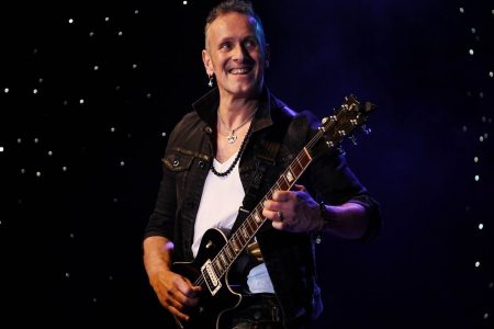 Def Leppard’s Vivian Campbell Says His Cancer Has Returned: “It Didn’t Stick” Def Leppard guitarist Vivian Campbell smiling on stage while playing a black Les Paul electric guitar under purple stage lighting