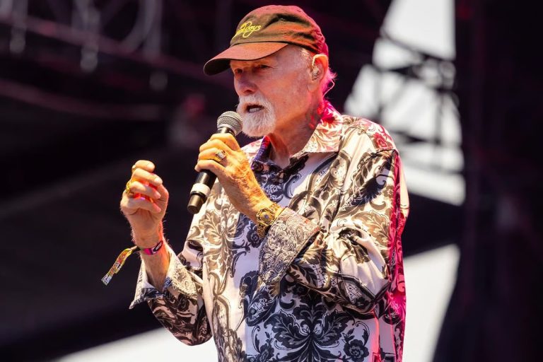 Mike Love of The Beach Boys singing into a microphone onstage in a Beach Boys cap and patterned shirt