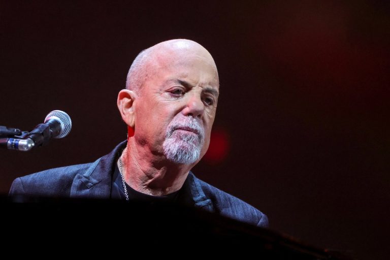 Billy Joel Reveals He Had Brain Surgery, and Why His Doctors Told Him to Stop Touring Billy Joel seated at a piano behind a microphone on a darkened concert stage, looking down with a reflective expression