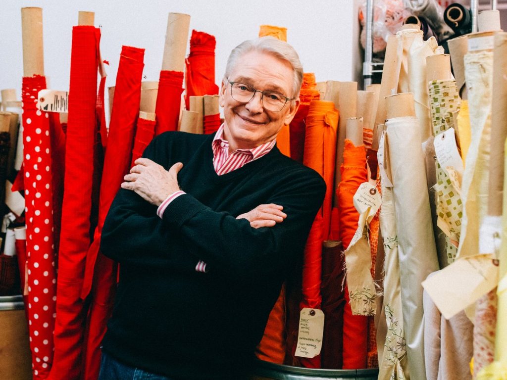 Costume designer Bob Mackie stands with his arms folded among tall bolts of red, orange and cream fabric in his studio
