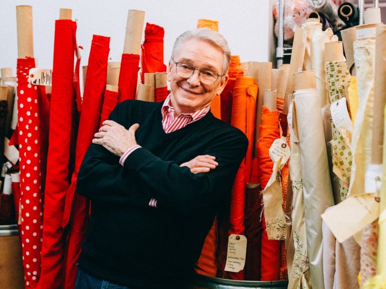 Costume designer Bob Mackie stands with his arms folded among tall bolts of red, orange and cream fabric in his studio